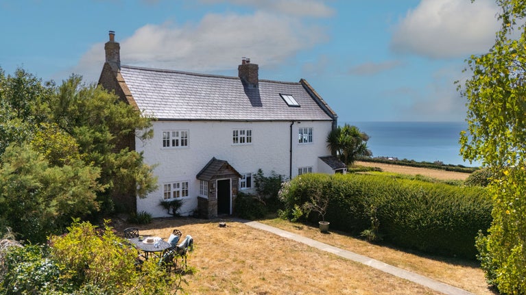 The exterior of semi-detached cottages, Labour in Vain Cottage and Chesil Cottage, with Labour in Vain's private garden in the foreground and the sea in the distance